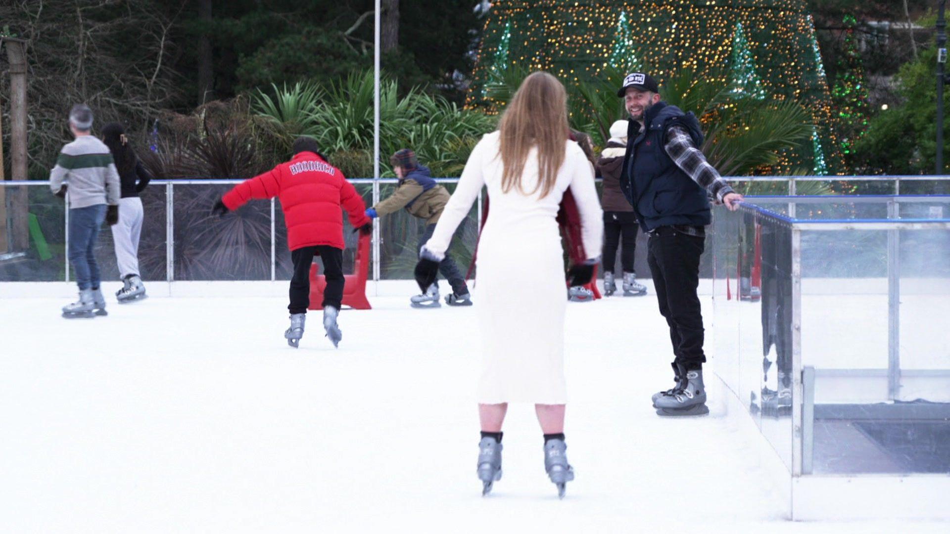 People on an outdoor ice rink. A woman, with her back to camera, wears a long white coat and ice skates. A man wearing skates and black clothing including a hat watches her whilst holding onto a rail. Other skaters can be seen in the background.