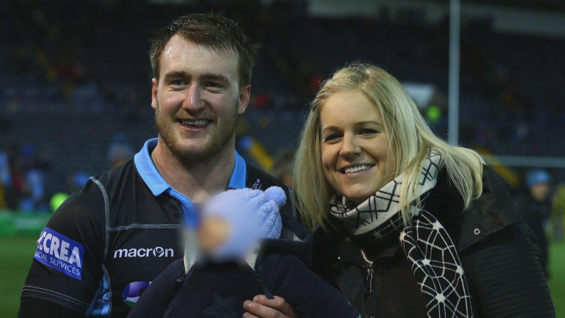 A man in a rugby top stands next to a woman with blonde hair both smiling on a rugby pitch and holding a child