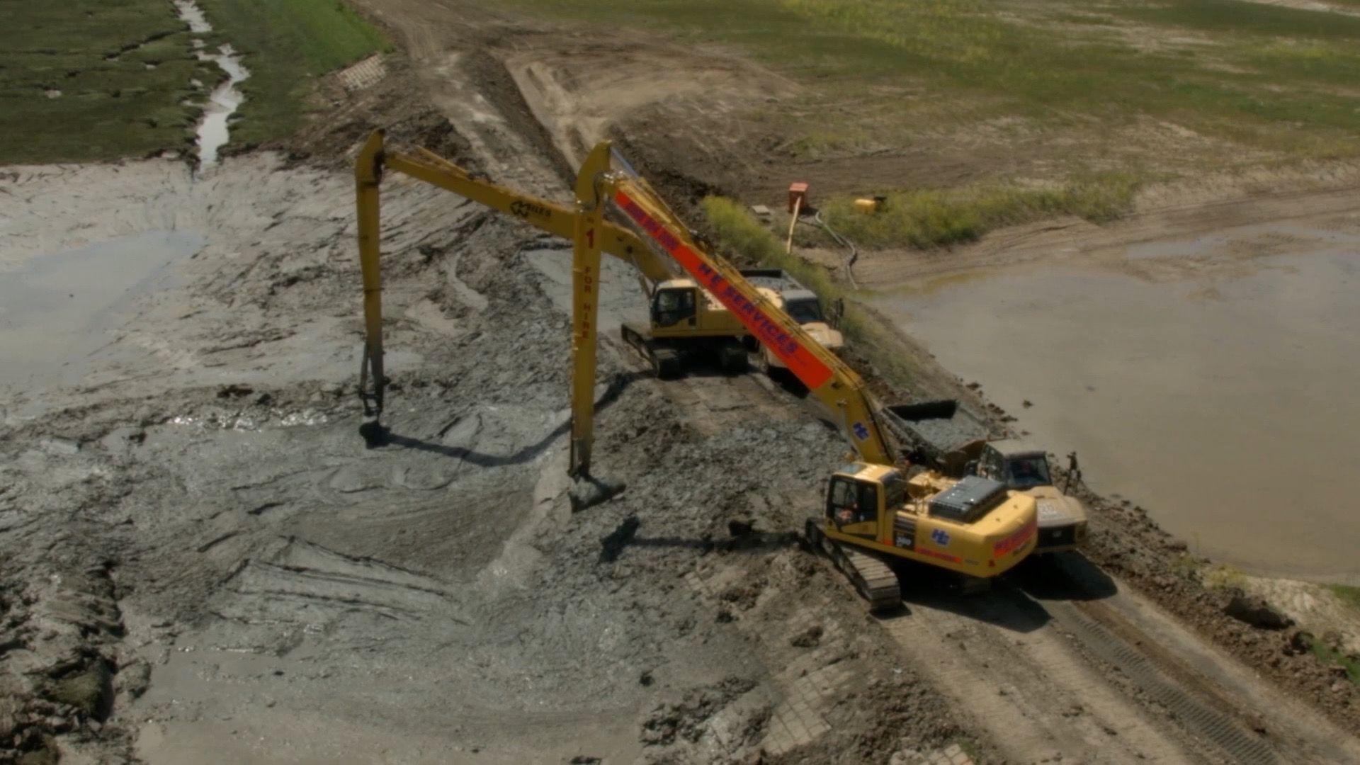 Two large yellow diggers work on lifting and shaping mud from trucks on a narrow road onto the wetlands next to it