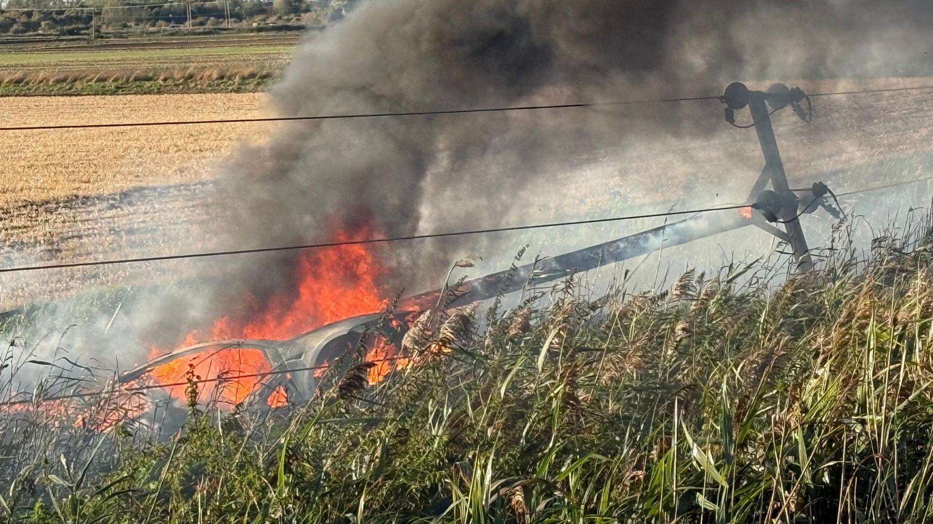 A vehicle is on fire in a ditch on the left with black smoke coming out of it. To the right is an electricity pole, which is black, and is in front of an grass ledge.