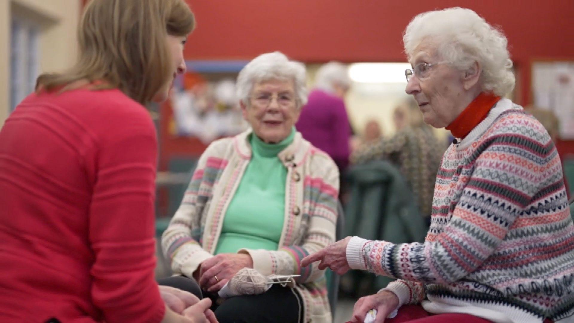 A group of three women sat in a circle. Younger woman wearing a red jumper chatting to two older women wearing knitted jumpers. One of the women is holding some knitting