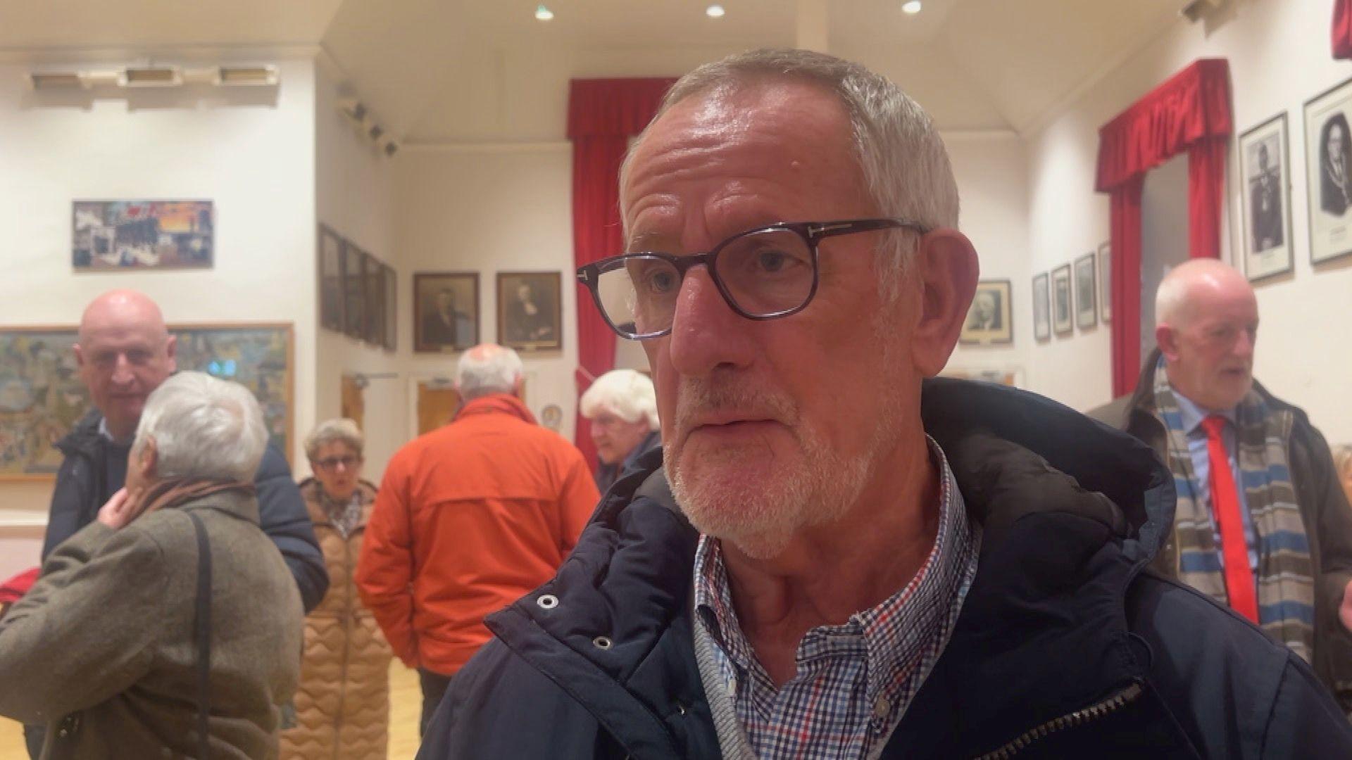 Lesley Norman is speaking in Grouville parish hall. There are five people behind him. He has glasses, white hair and a beard. 