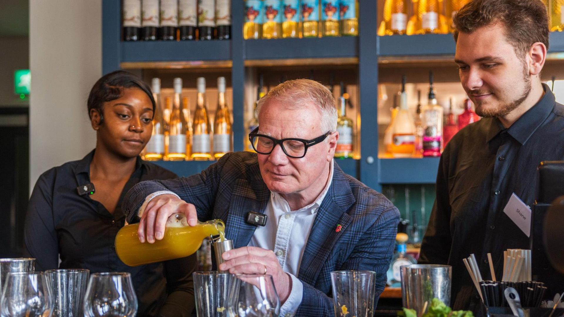A man with silver hair, black glasses and a blue checked suit pours an orange liquid from a glass bottle into a cocktail mixer. A young woman and man stand either side of him, all of them behind a bar with bottles lined up on blue shelves behind them.