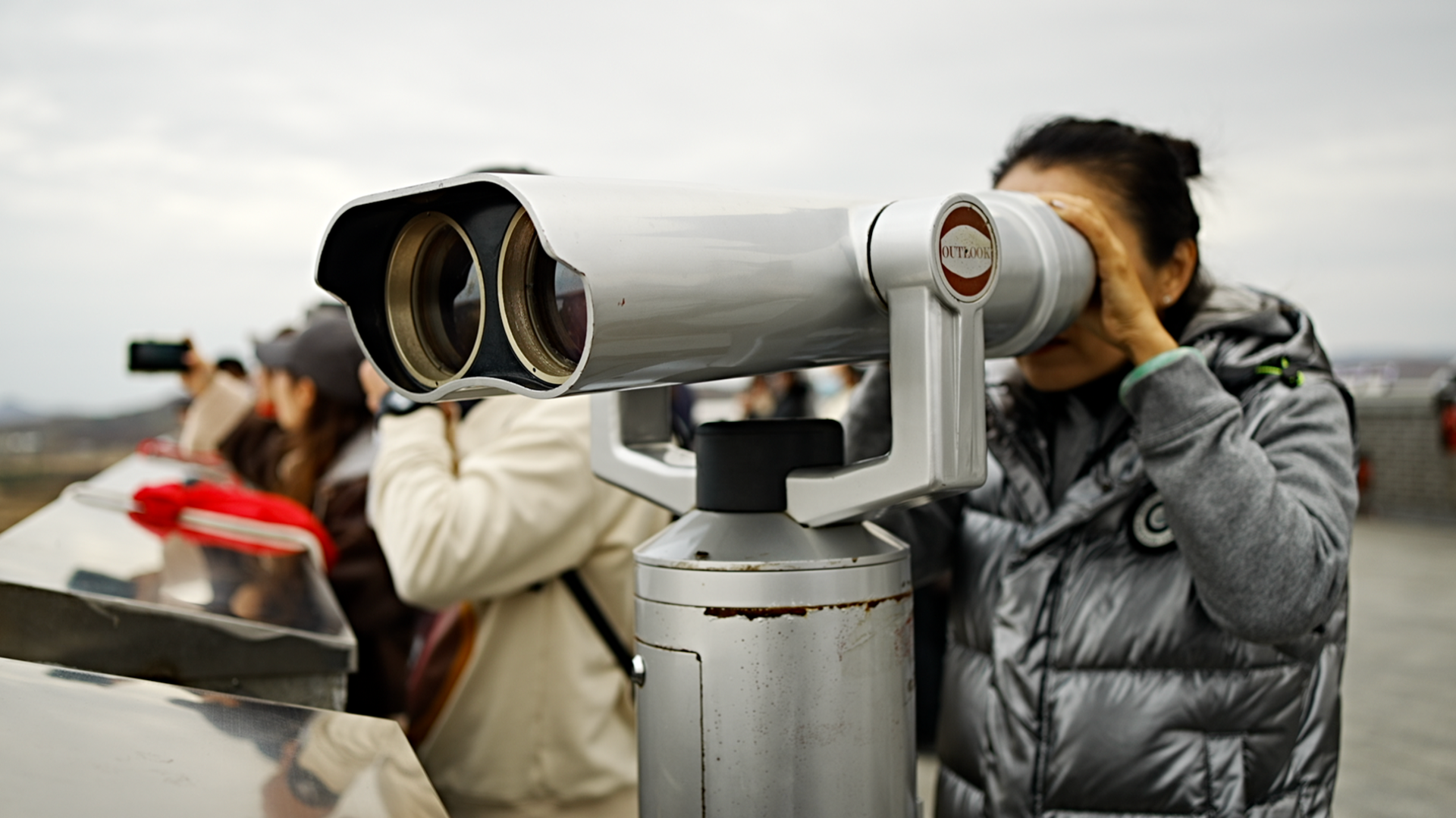 Chinese tourists wrapped in warm coats in Fangchuan look across the border using large grey binoculars under a cloudy sky