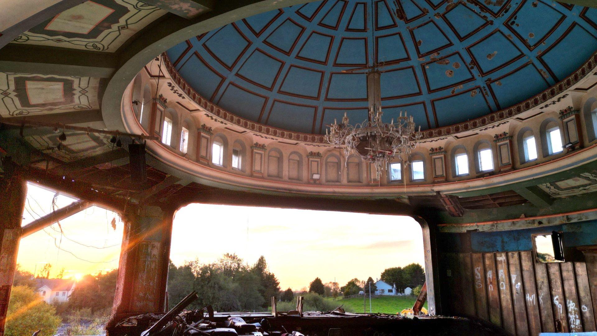 Edendork Hall from the inside after demolition - the inside of the dome is still intact, it's painted white frame with blue in the dome and gold linbing. There is a chandelier still intact covered in cobwebs/rubble. From the open section you can see out to trees, houses and the sunset which is reflecting back into the partial building. 