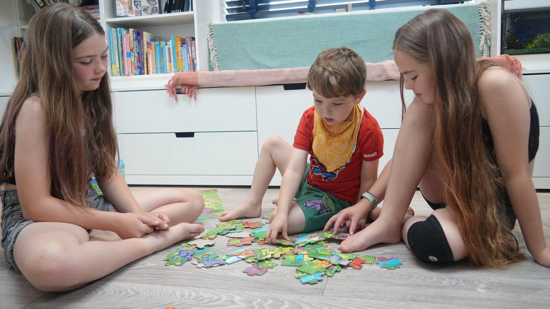 Bobby sits on the laminate floor doing a jigsaw with his two sisters, who are sitting cross-legged either side of him. They both have very long wavy dark blonde hair and wear shorts and T-shirts. Bobby has short wavy hair and wears green shorts and a red T-shirt. Behind them is a white storage unit with drawers and shelves.