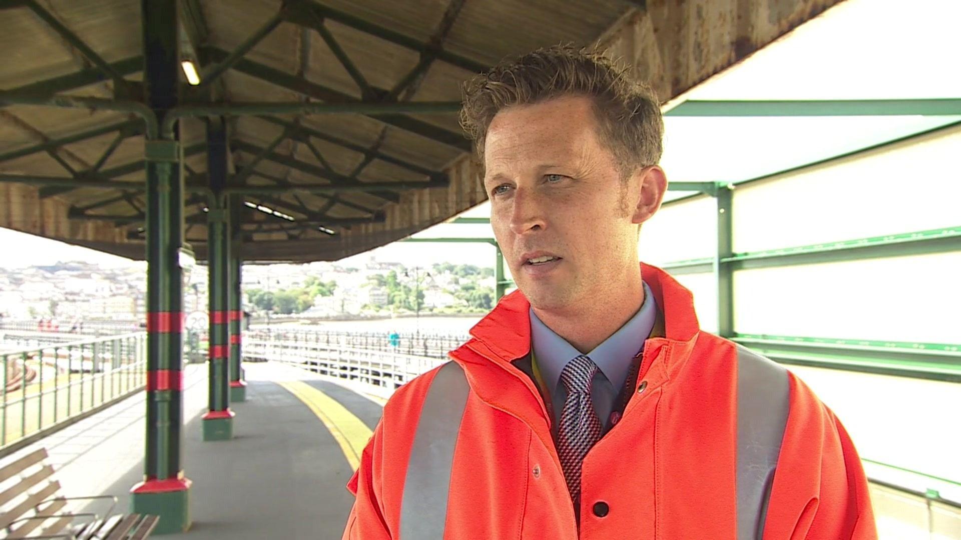 A man wearing a shirt and tie with an orange high-visibility jacket standing on a train platform with the railway behind him.