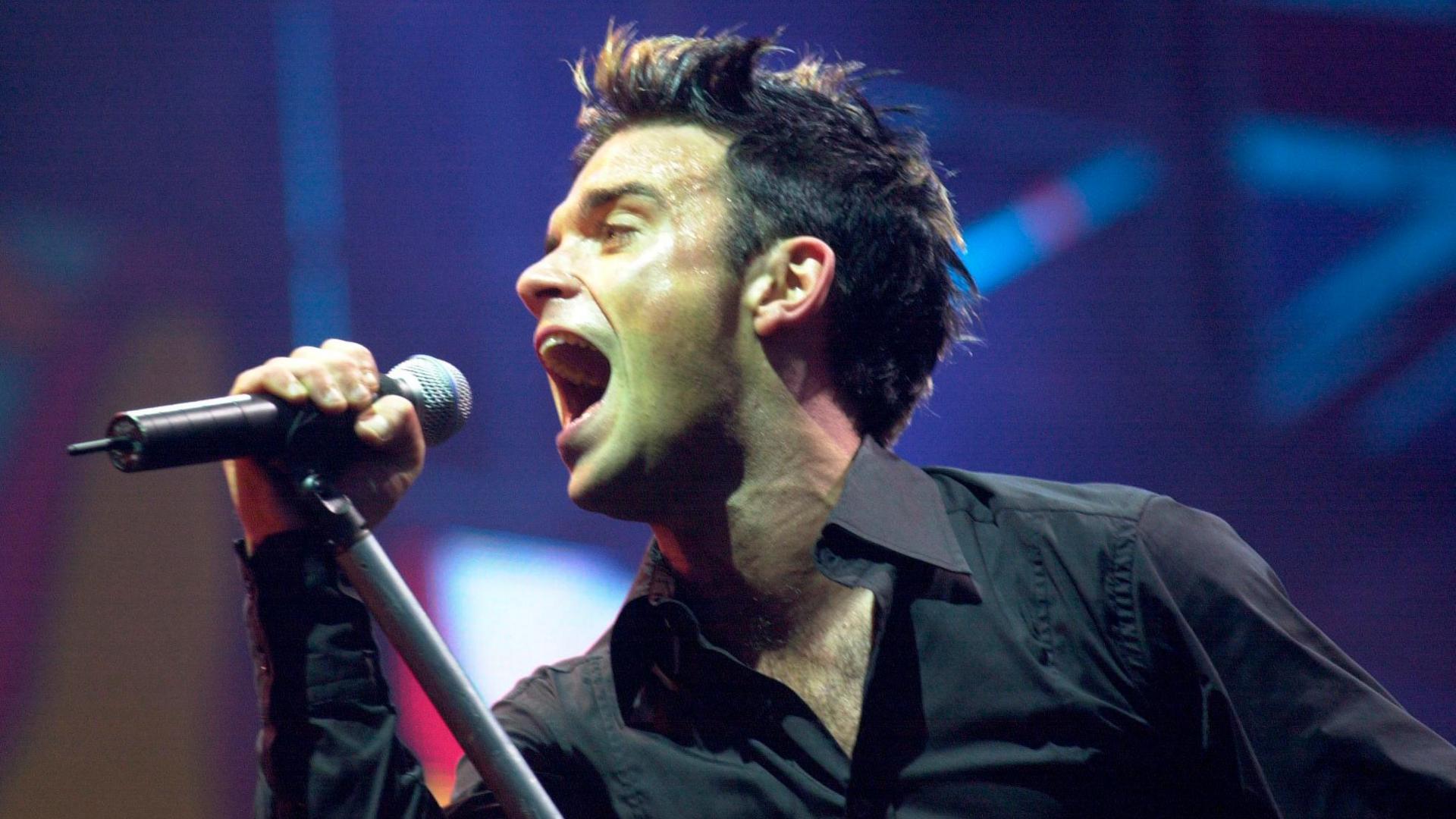 Robbie Williams singing on stage in 2001 at Milton Keynes Bowl. He has blonde highlights in his dark hair and is holding the microphone as he sings into it.