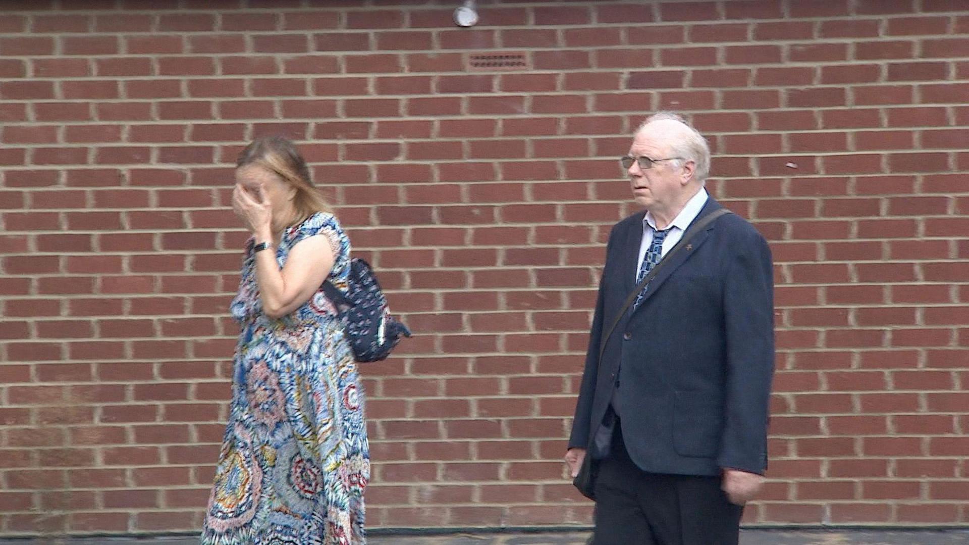 David Eggleton wearing a dark blue suit and walking into court with his arms hanging down by his sides. He is joined by a woman in a patterned dress who is covering her face