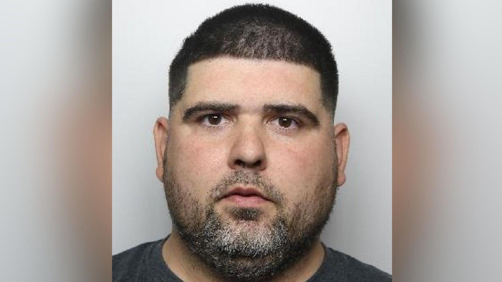 A mugshot of Ackerman who has short black hair, brown eyes and a facial hair. He wears a grey t-shirt as he looks at the camera unsmiling.