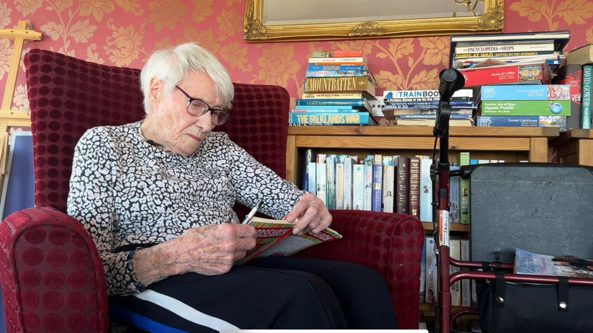 A woman sits in an armchair in front of a bookshelf. She is reading a crossword puzzle book.