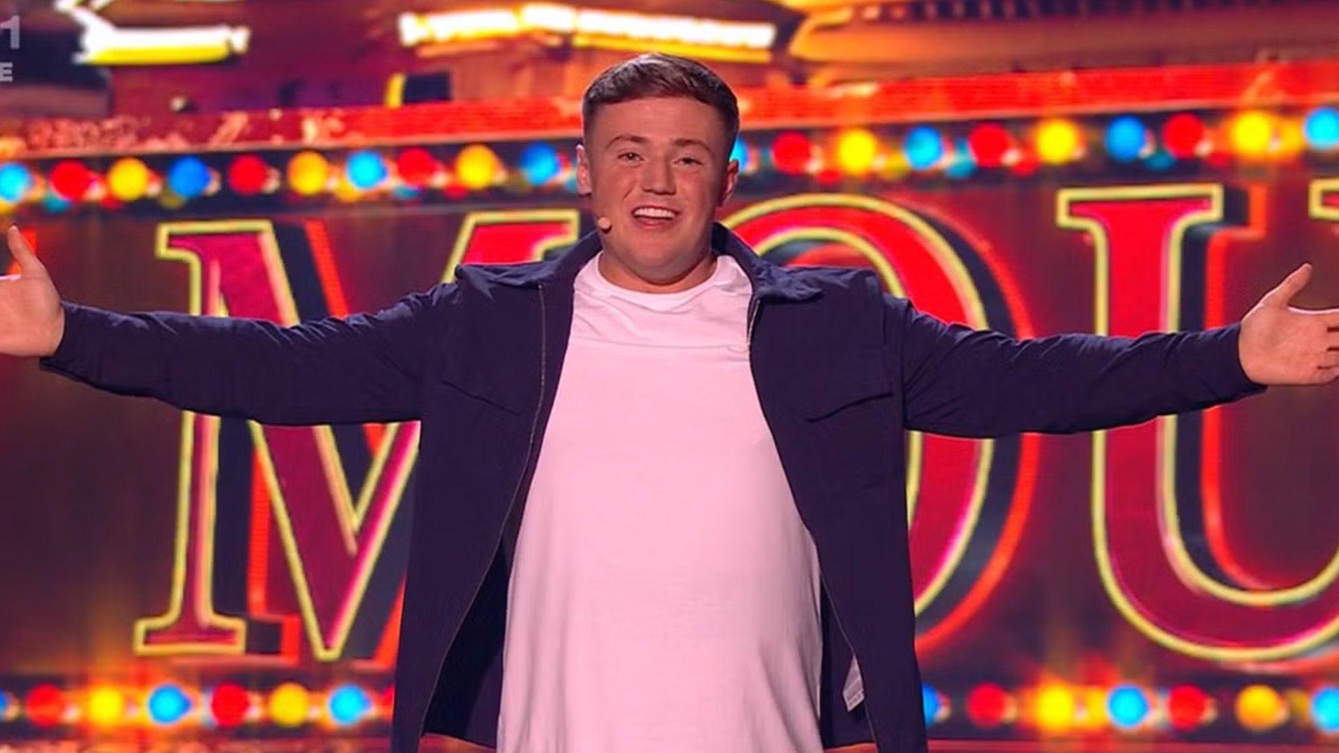 Harry Moulding stands on the stage looking down the camera with his arms outstretched.