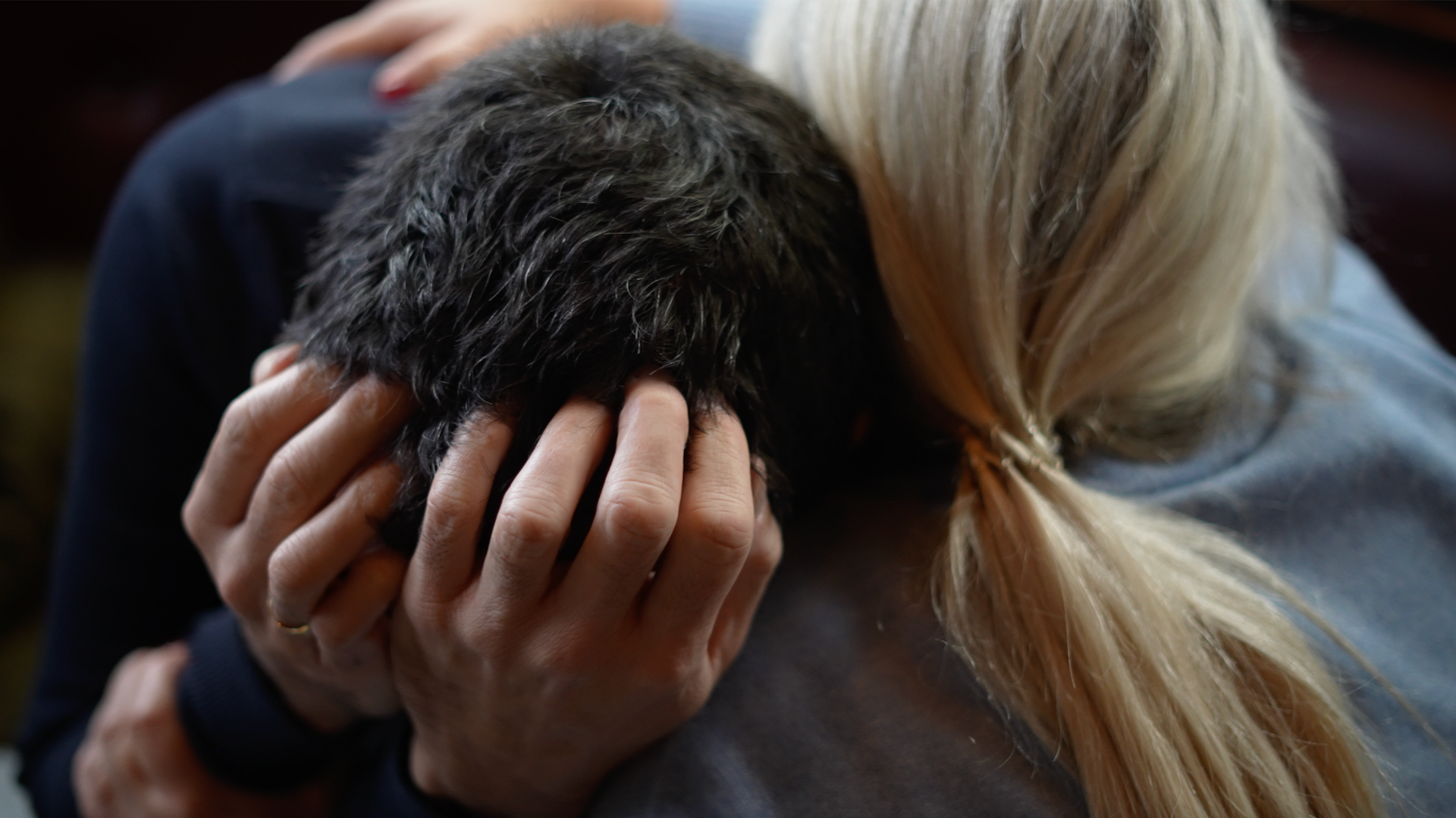 A man and woman embrace. The man, with dark hair, has his head in his hands and the woman, with blonde tied back hair is holding him. 