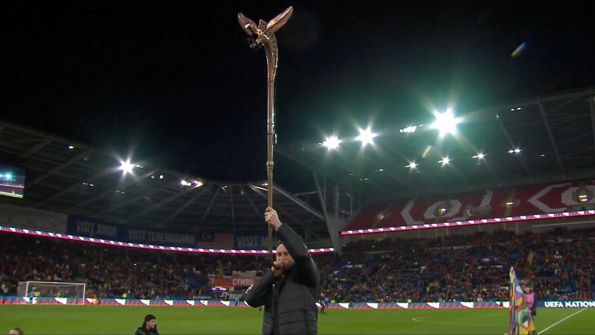 A man stands on a football pitch before thousands of fans, playing a long, bronze horn.