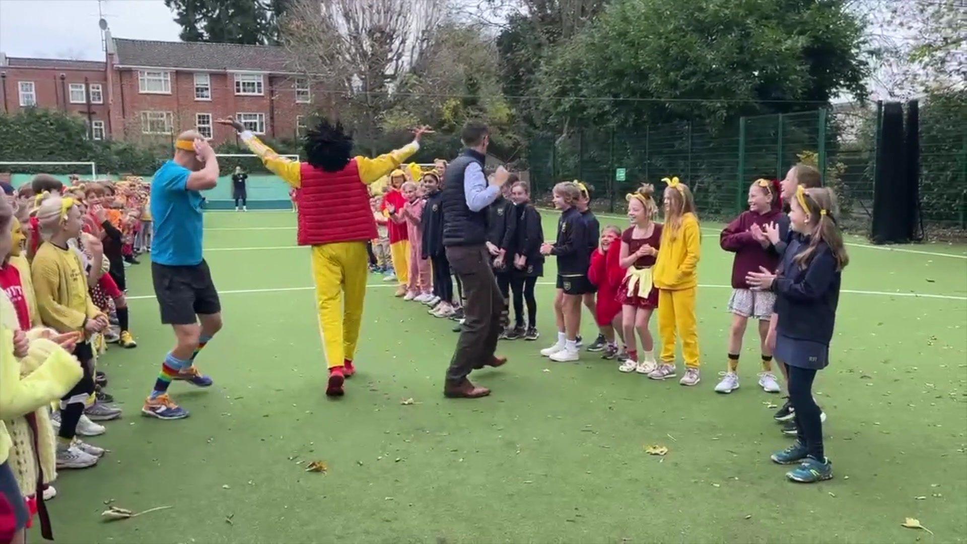 Adults running between cheering children. All are wearing Pudsey style yellow ears and stood on a green astroturf 