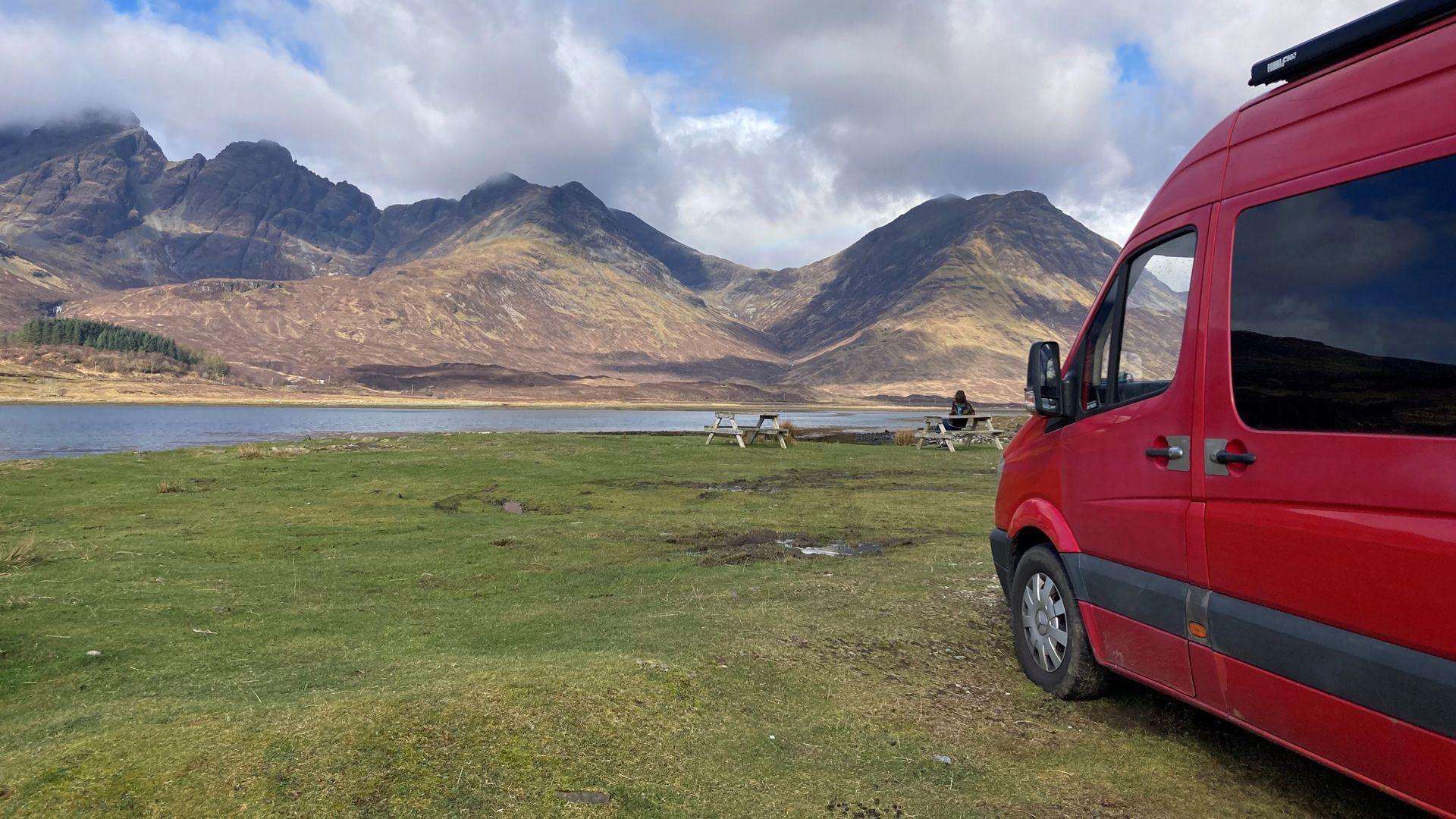 A red campervan parked next to a beautiful lake