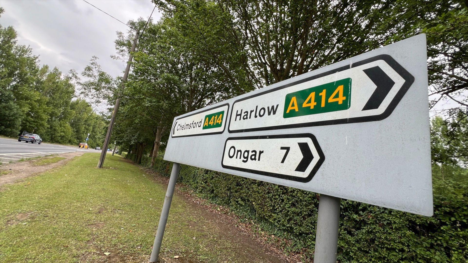 A road sign which shows Chelmsford in the left direction (with yellow writing on a green backdrop saying A414), Harlow in the right direction (with yellow writing on a green backdrop saying A414) as well as Ongar in the right direction, and a number 7 indicating it is seven miles away. The sign is in front of a hedgerow and trees, with a road running along the left-hand side of screen.