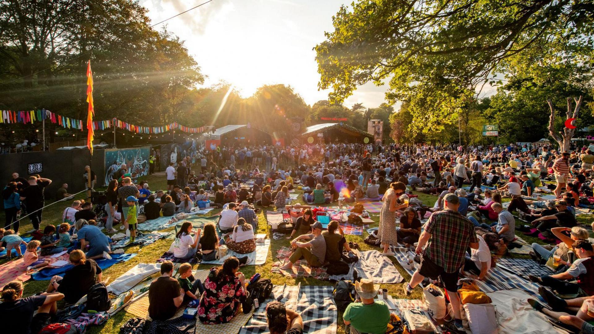 A large number of people sat on the grass with stalls in the background