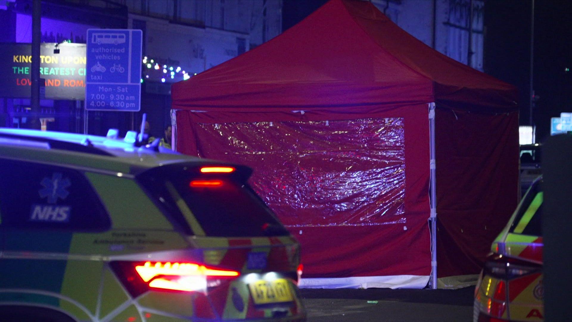 A red police tent is set up in the road where the incident took place.  Two emergency vehicles are parked in front of the tent.  The picture is taken in the dark.