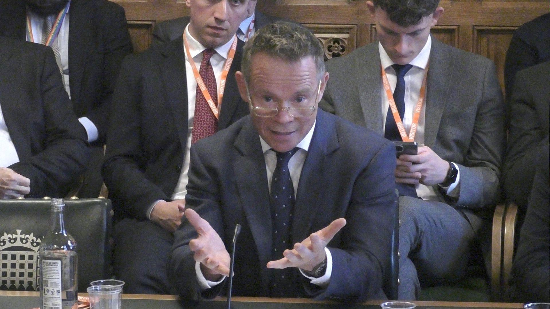 Tom Keith-Roach, centre frame and part way through his evidence to a House of Commons select committee. He is mid-flow, with his mouth partly open and both hands spread in front of him. His is wearing glasses that are part way down his nose and he is looking over them. His eyes are wide and his brow furrowed. He is wearing a navy suit jacket, blue tie and white shirt. He is sitting at a table, whose green leather top can just been seen at the bottom of frame. An empty chair on the left of frame is dark green and bears the Parliament crowned portcullis logo. Behind Mr Keith-Roach are several people, also wearing suits and lanyards with 'visitor' written on them. One of the people behind is checking their phone. Behind those people a wood-panelled wall can just be seen. 