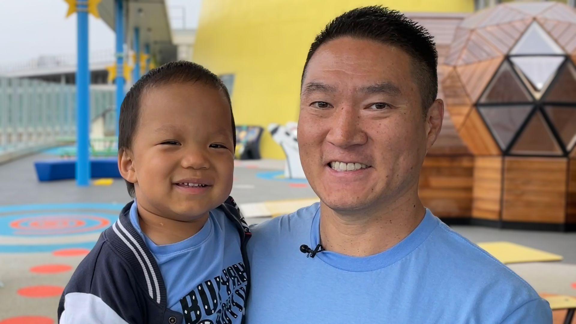 Dad Ricky is carrying Oliver in his arms.  They are borth smiling and wearing blue T-shirts. They are on a rooftop with colourful seats and tables in the background.
