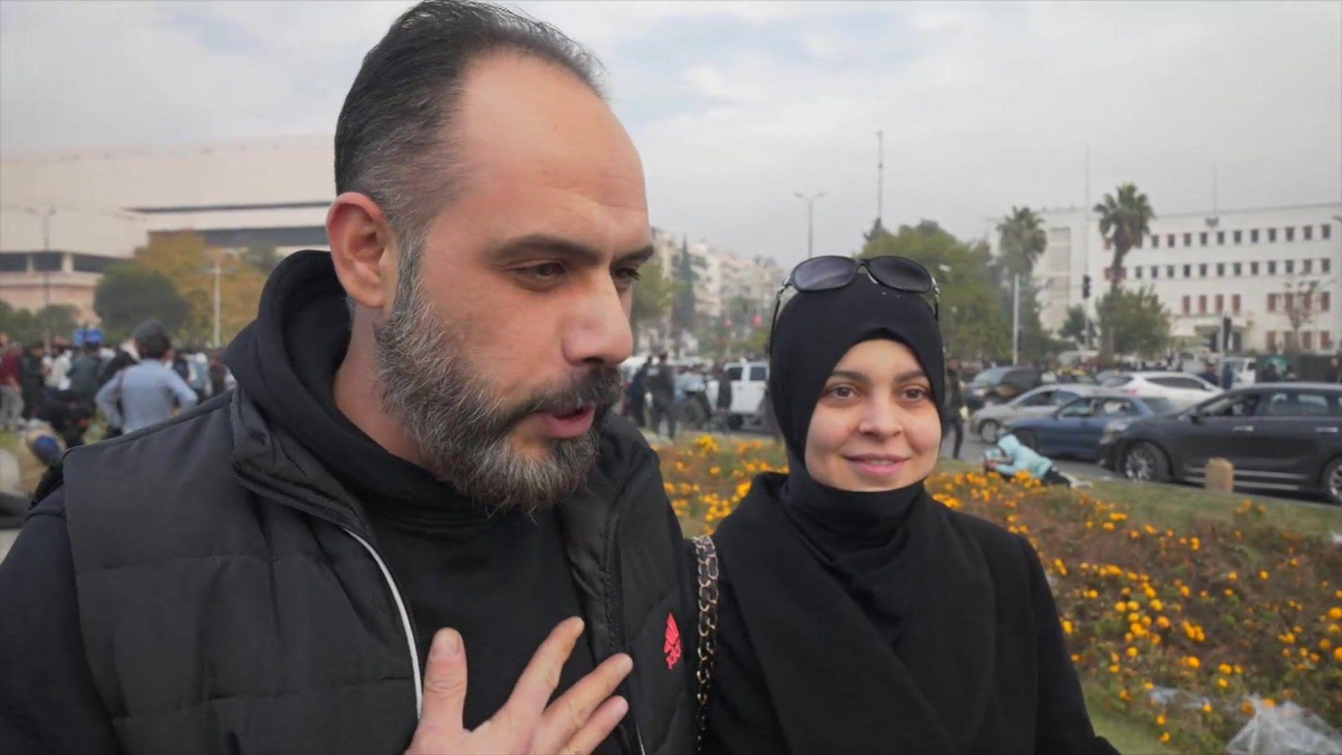 A Syrian man is seen standing on a street with his wife being interviewed by a reporter. He has his hand to his chest as he speaks. He is wearing a black hoodie. 