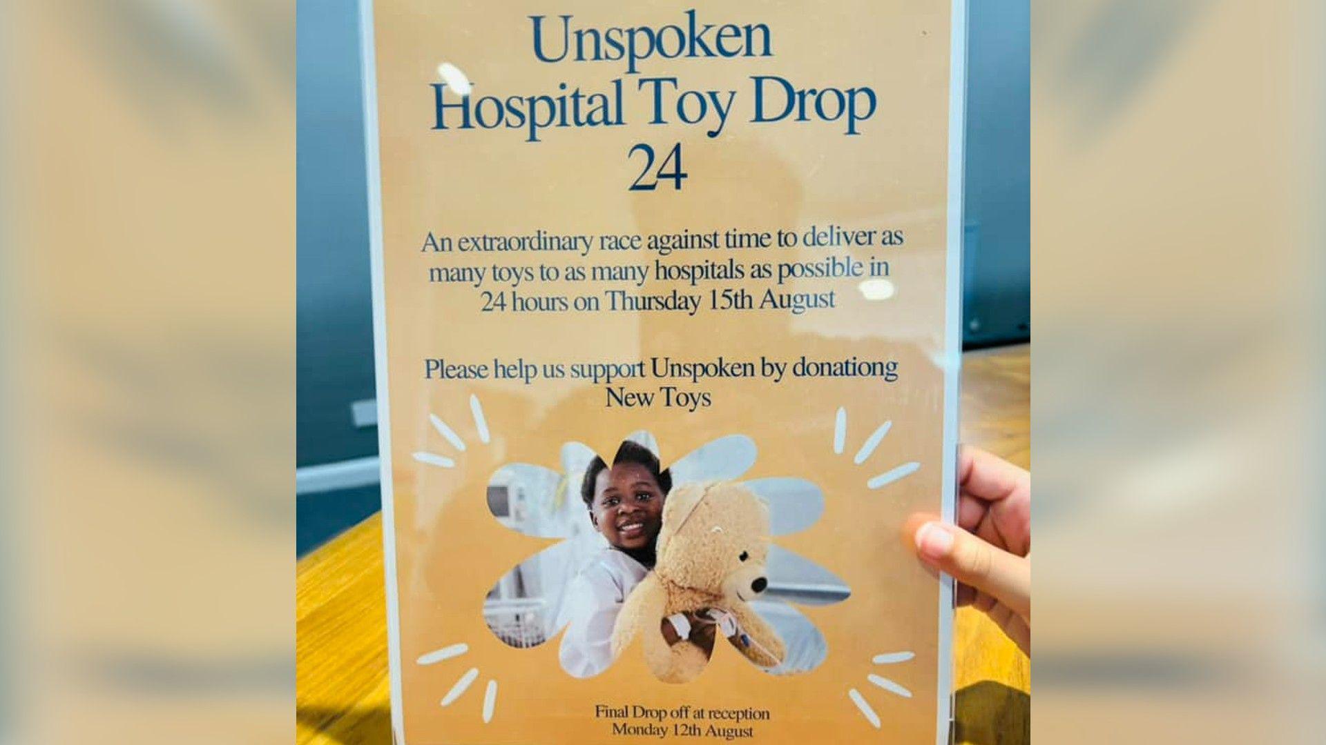 A hand holding up a poster advertising the drop off point, which reads "Unspoken Hospital Toy Drop 24", with a photo of a child holding a teddy bear