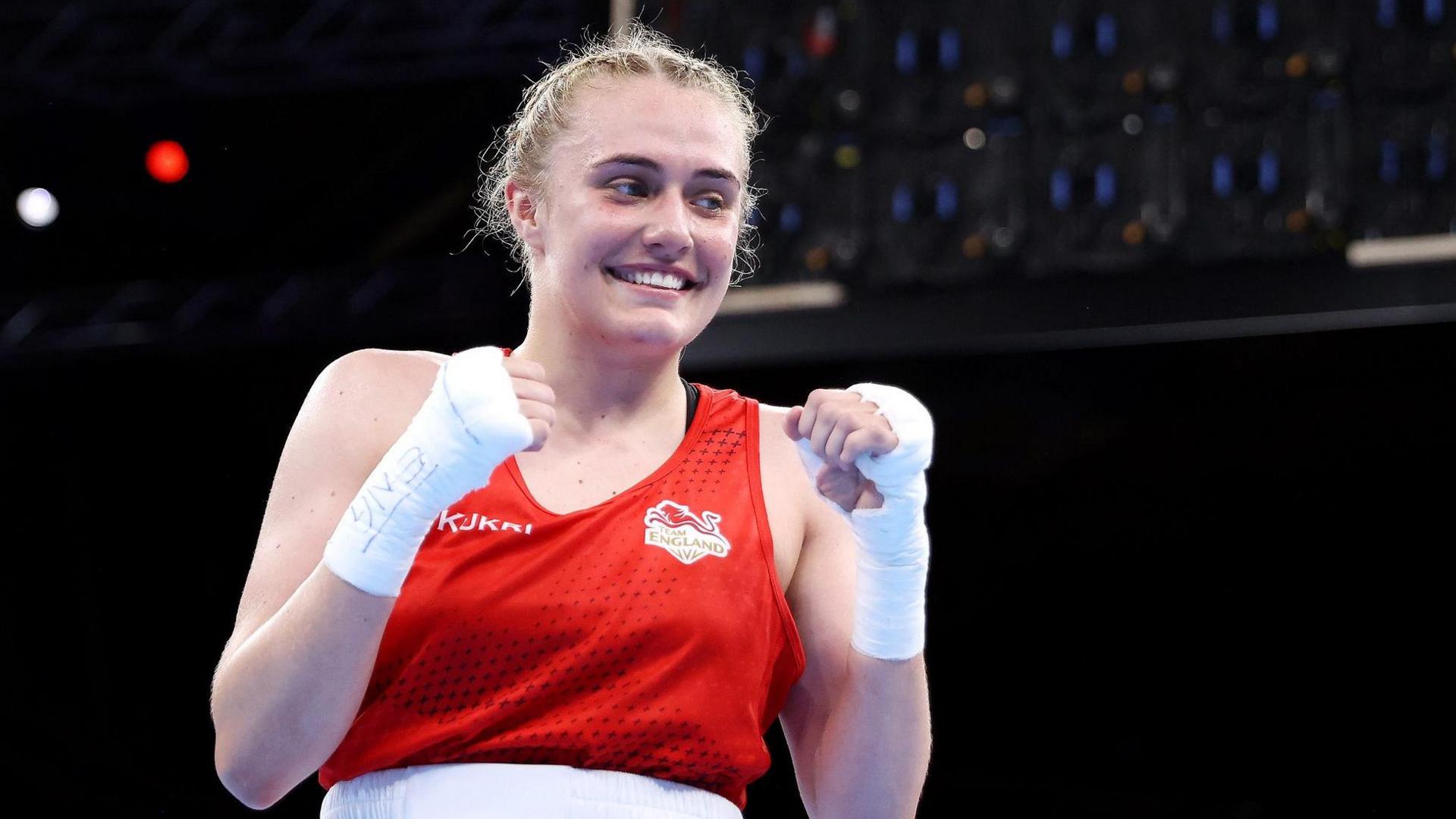 Gemma Richardson in red England boxing kit, her hands are wrapped and she is making fists and smiling. Her blonde hair is plaited close to her head.