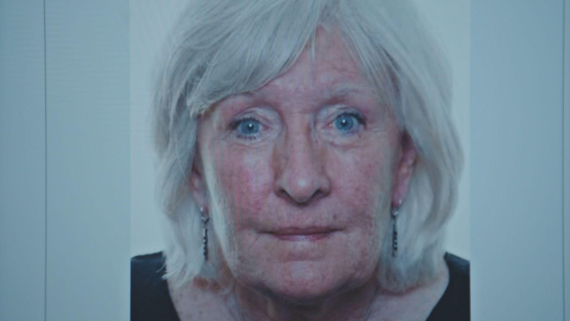 A close up shot of Annette as she stares at the camera with her eyebrows slightly raised. She has bright blue eyes and smartly coiffed white hair. She wears dangly silver earrings