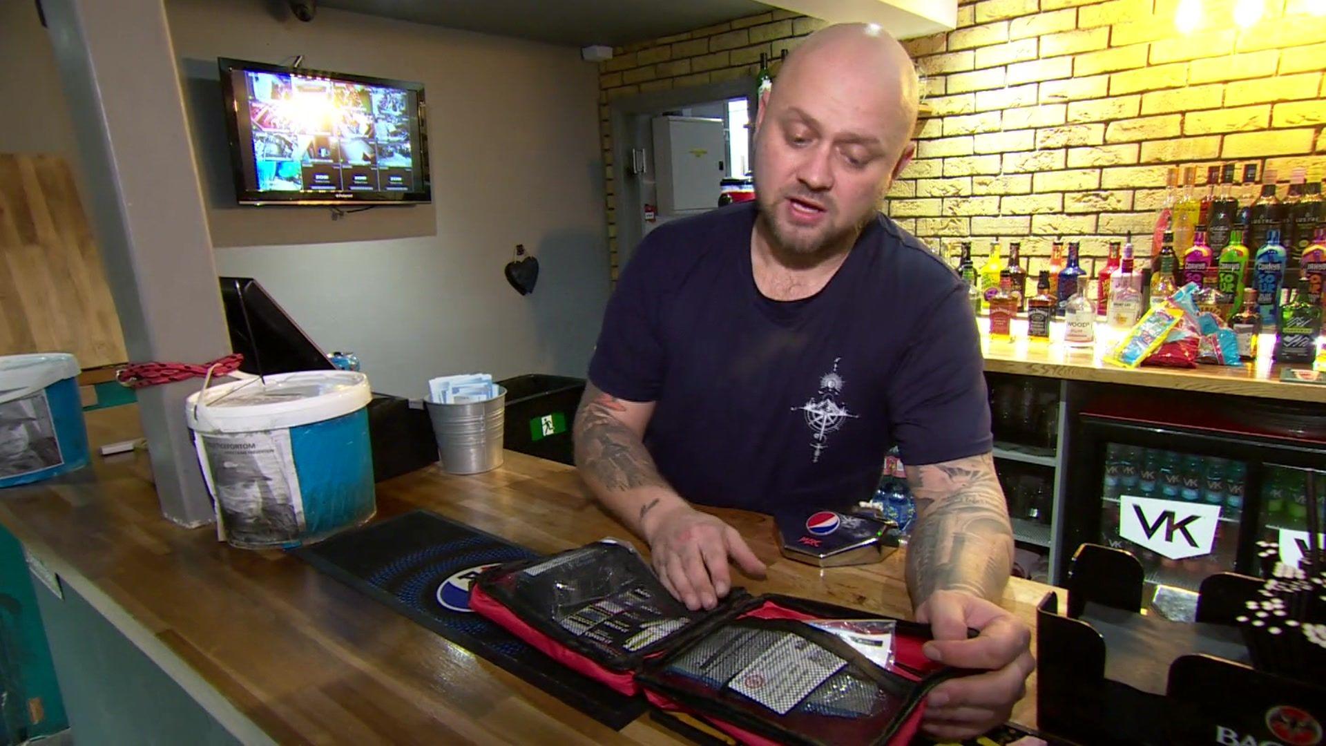 Daniel Cooke, who wears a blue T-shirt and has tattoos on his arms, stands behind the bar of a club and demonstrates the contents of a bleed kit - a package of bandages, trauma dressings and other first aid equipment - housed in portable a red bag.