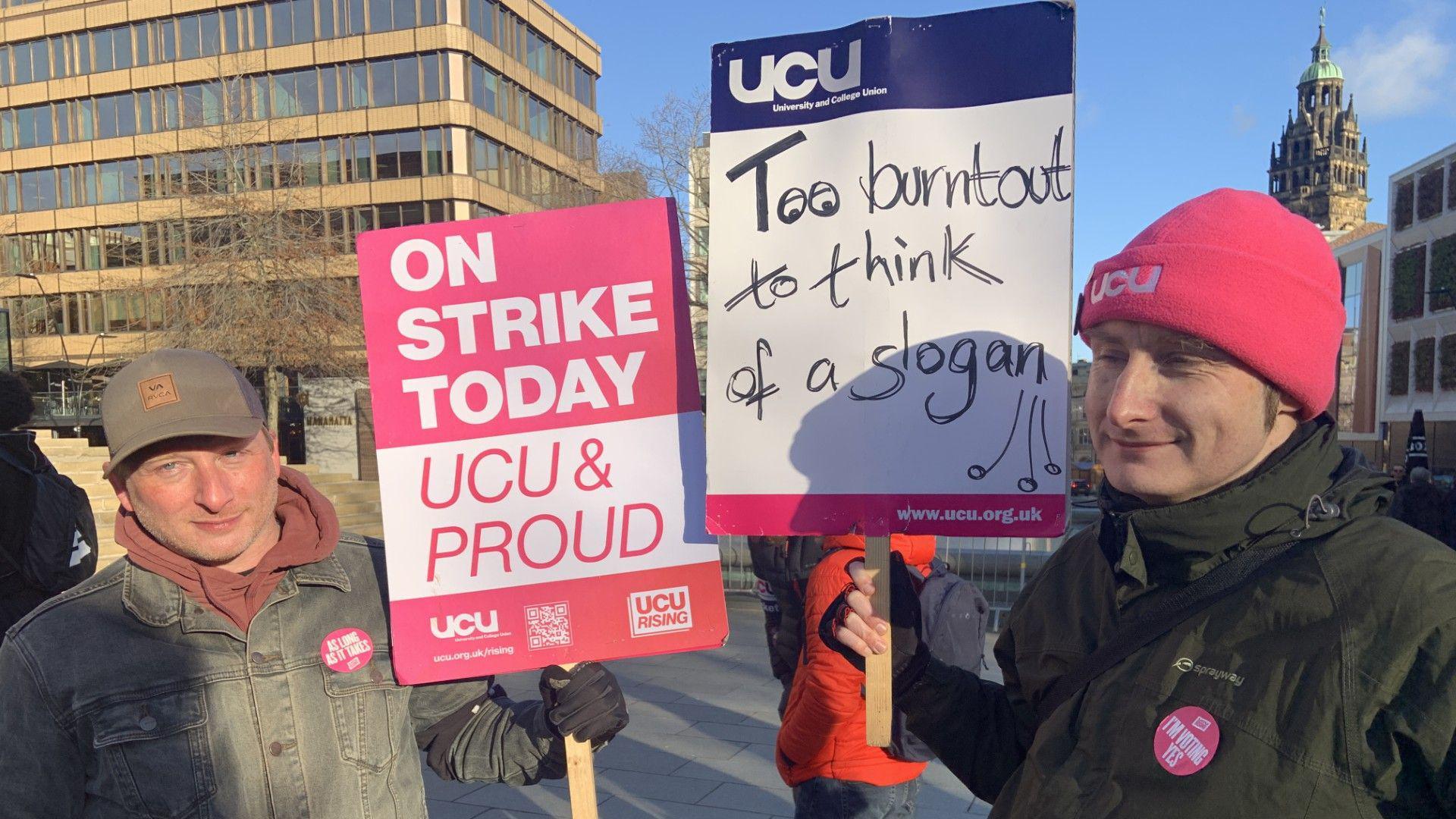 Two men are standing in the city centre. One man in a baseball cap and denim jacket has a placard in pink and white saying Strike Today UCU and Proud. A man next to him has a pink beanie hat and green coat and is holding a placard saying Too Burnout To Think Of A Slogan