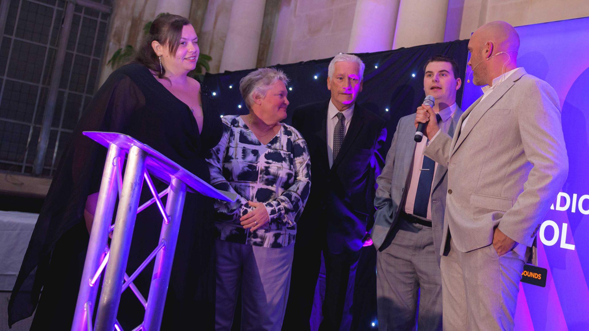 Two women and two men in smart outfits are onstage answering questions on a microphone held by presenter Joe Sims. He is wearing a grey suit with a white shirt.