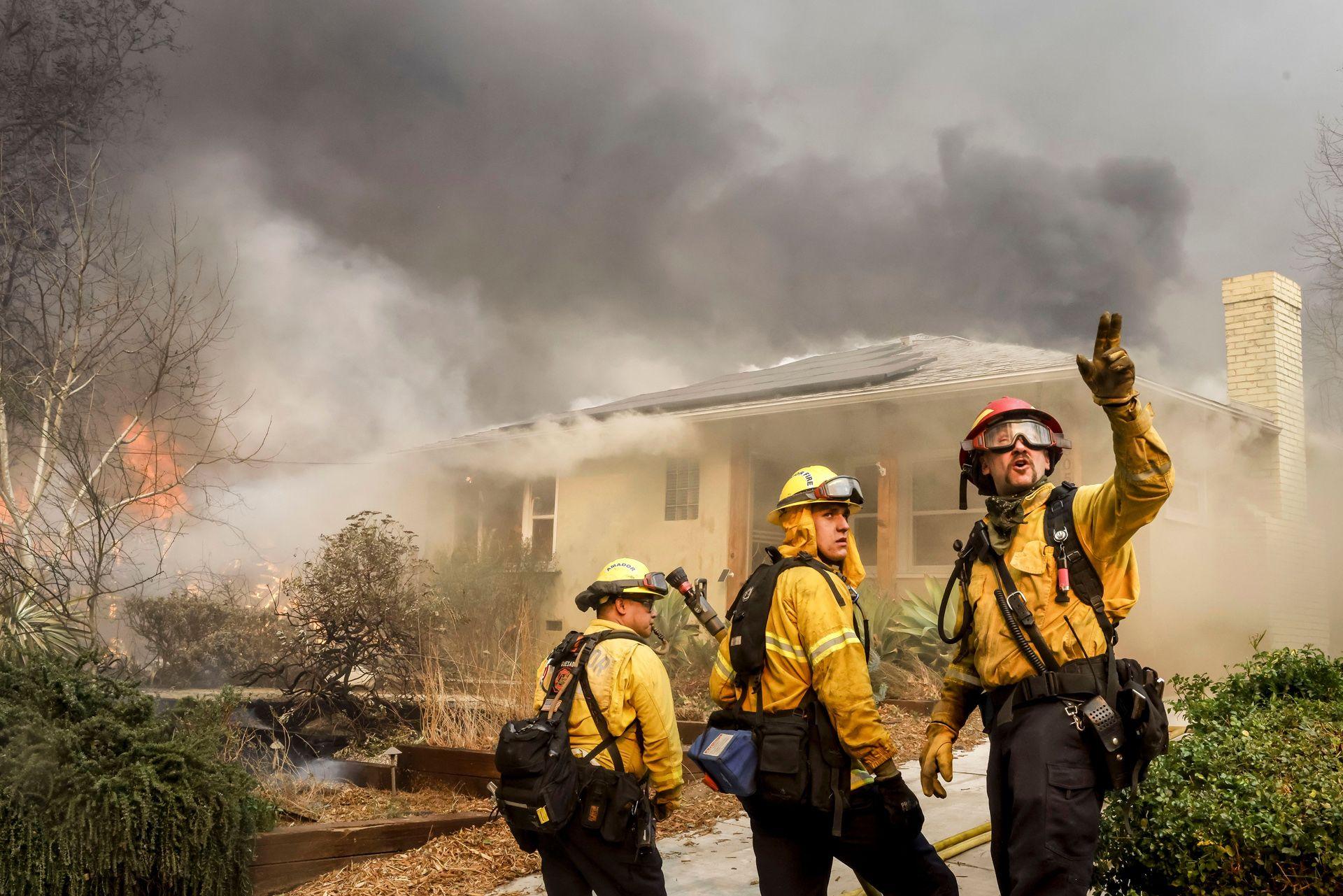 Three Los Angeles County firefighters look away from camera and gesture next to a burning home while trying to protect homes from the Eaton wildfire in Altadena on Wednesday