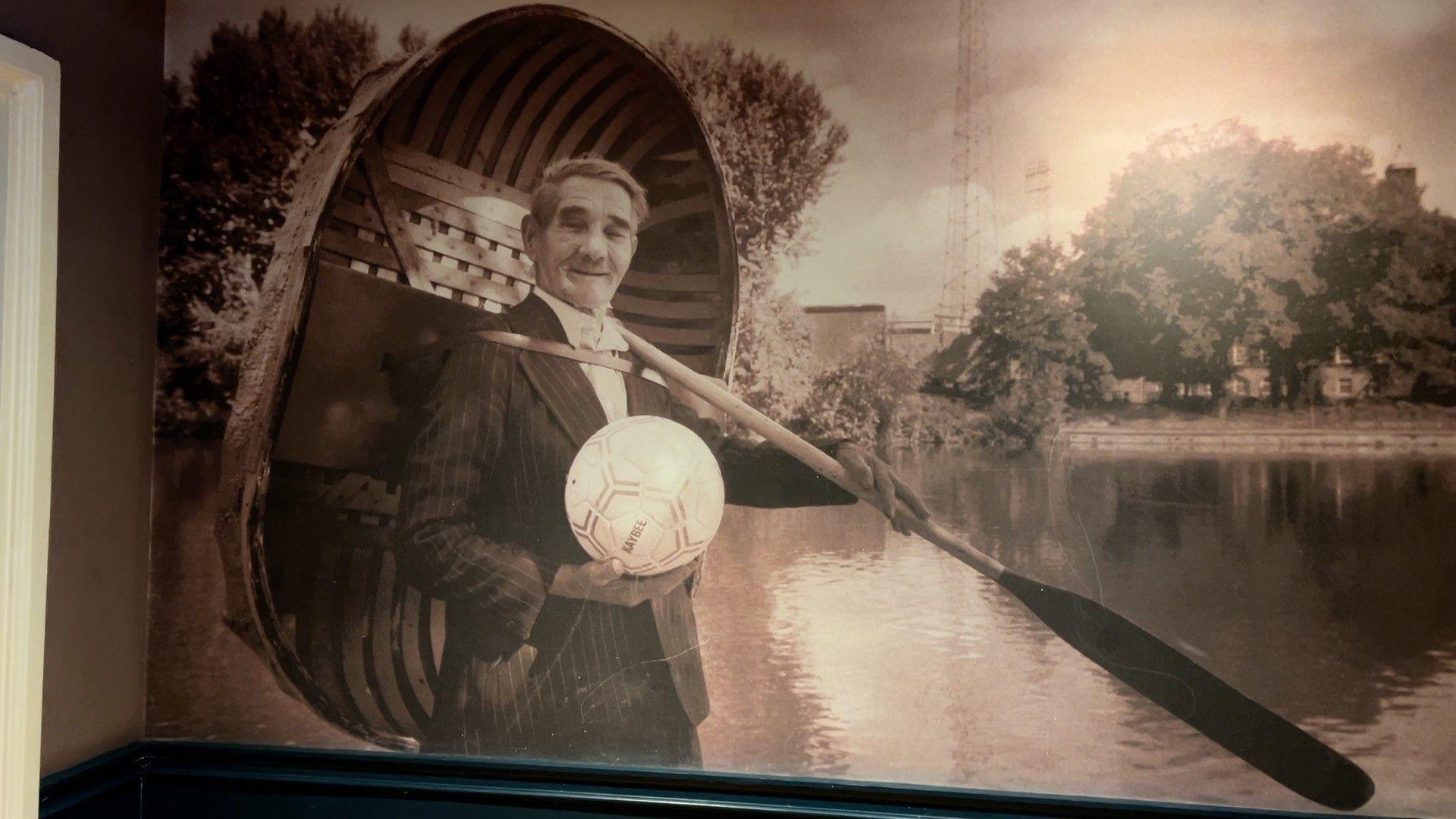 A large sepia-toned photograph on a wall. The photo is of an older man with a pin-striped suit on. He is holding a wooden coracle on his back that is circular and has a small bench in it. He also holds a wooden oar, and a white football in his hand.