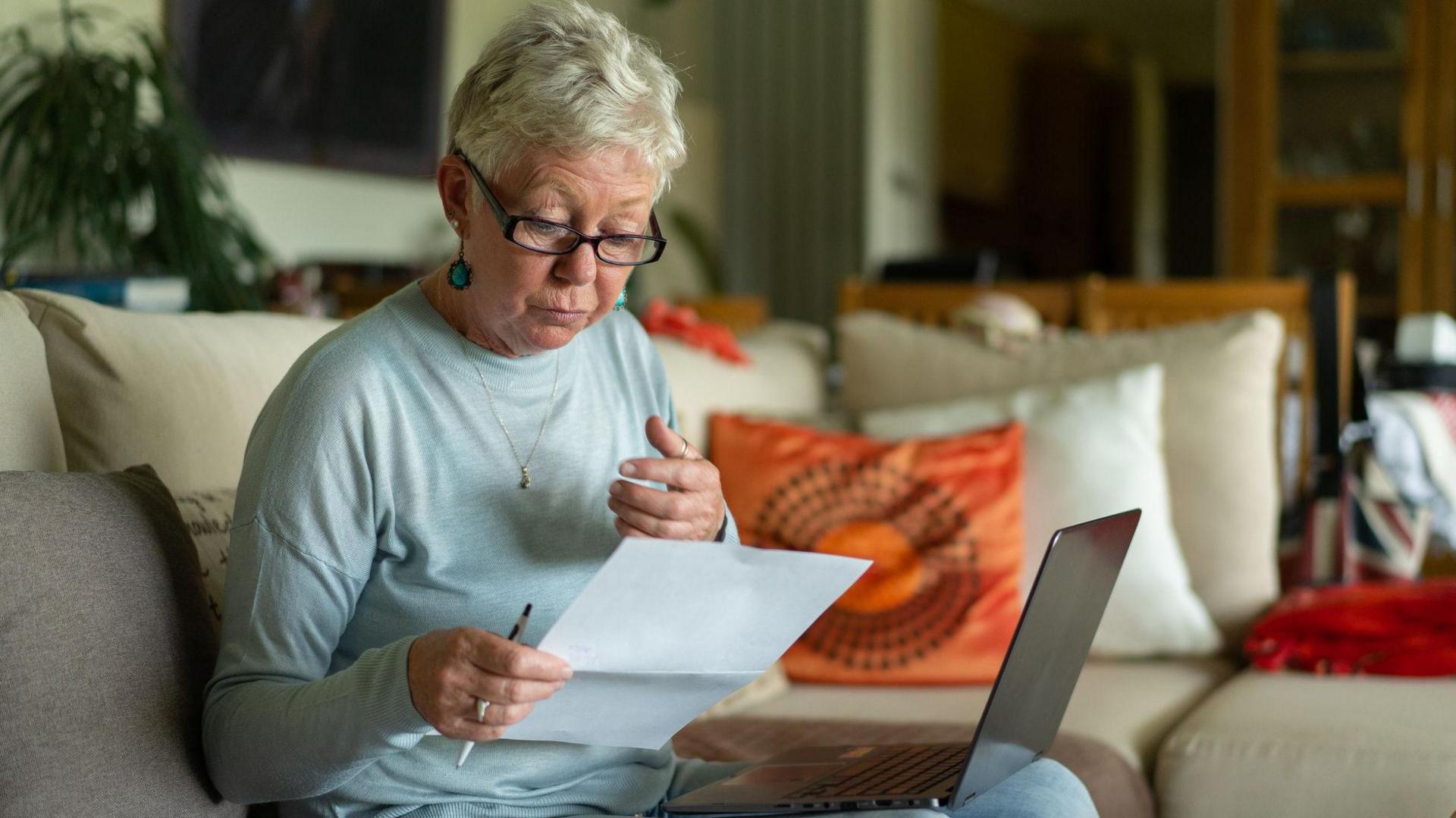 An elderly lady looks at a bill while sitting with a laptop on her knee on a sofa