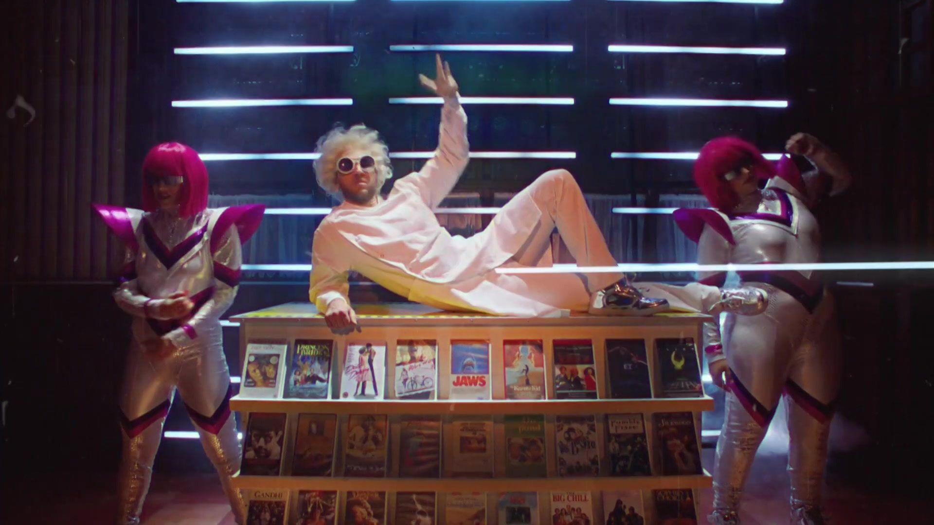 A man wearing a blonde bushy wig, sunglasses, white trousers and jacket lies across a stand containing copies of films from the 70s and 80s. Women in silver space age-style suits stand either site of him wearing pink wigs and sunglasses.