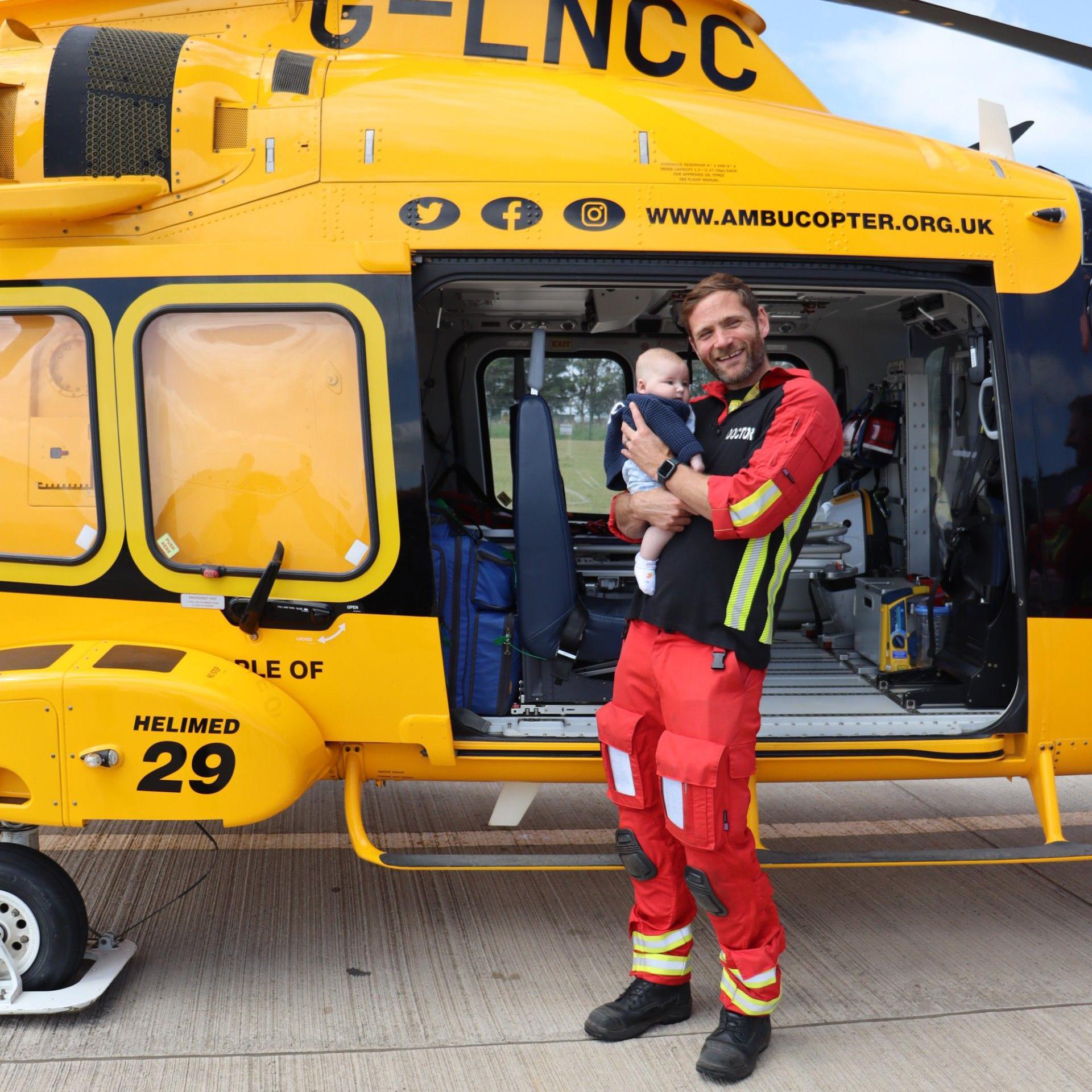 A doctor wearing a red and black uniform, who has short brown hair and a beard, holds a baby boy in his arms as he stands in front of a yellow air ambulance helicopter on the runway of an airfield.