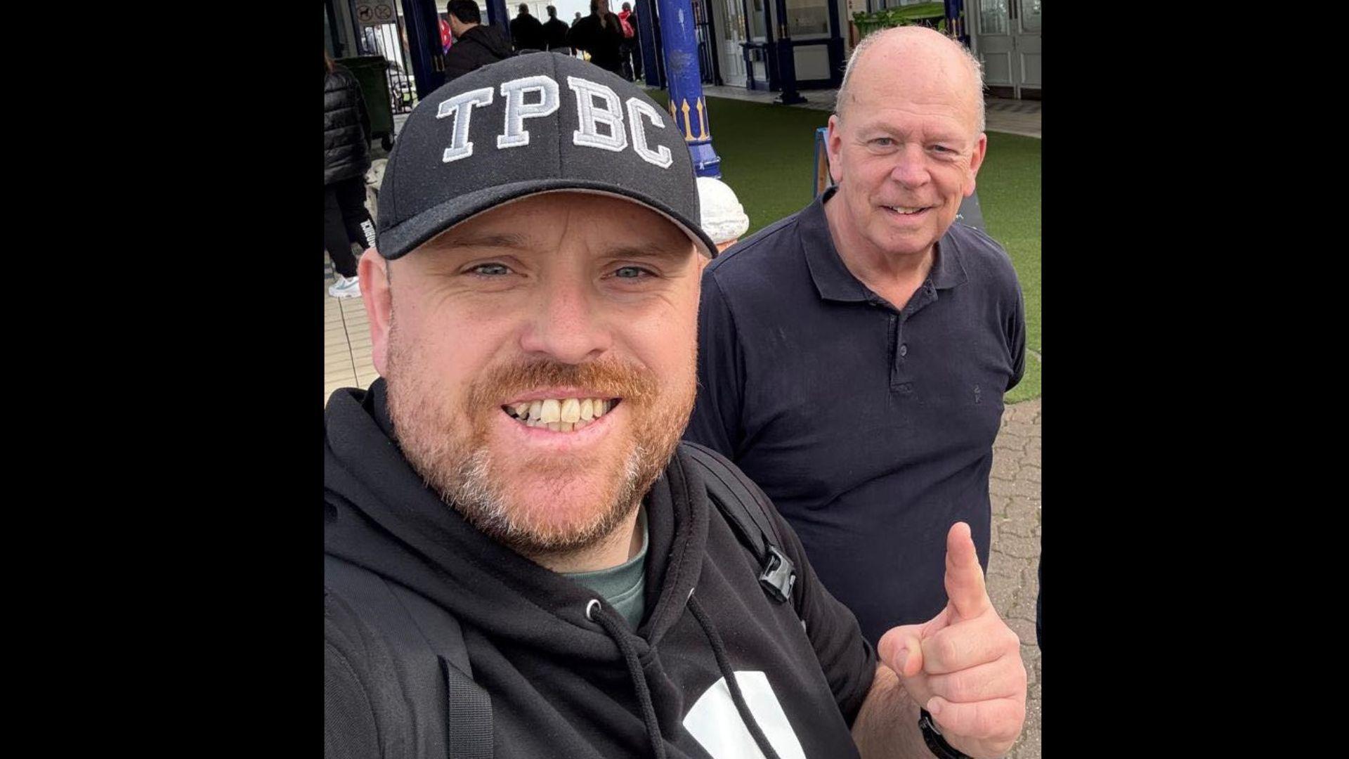 A man in a black hoodie and black hat, and a man in a black polo shirt are standing in front of Eastbourne Pier.