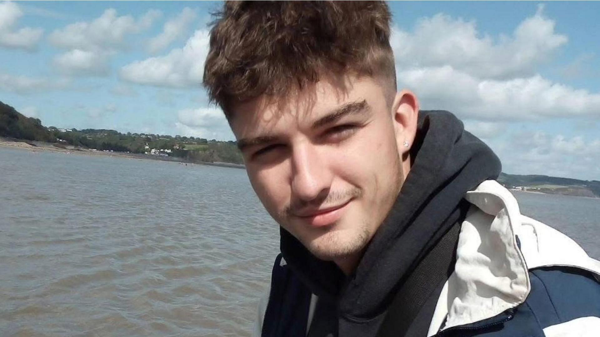 A young man with short dark hair, with a black scarf around his neck and a white jacket. He is standing in front of a body of water.