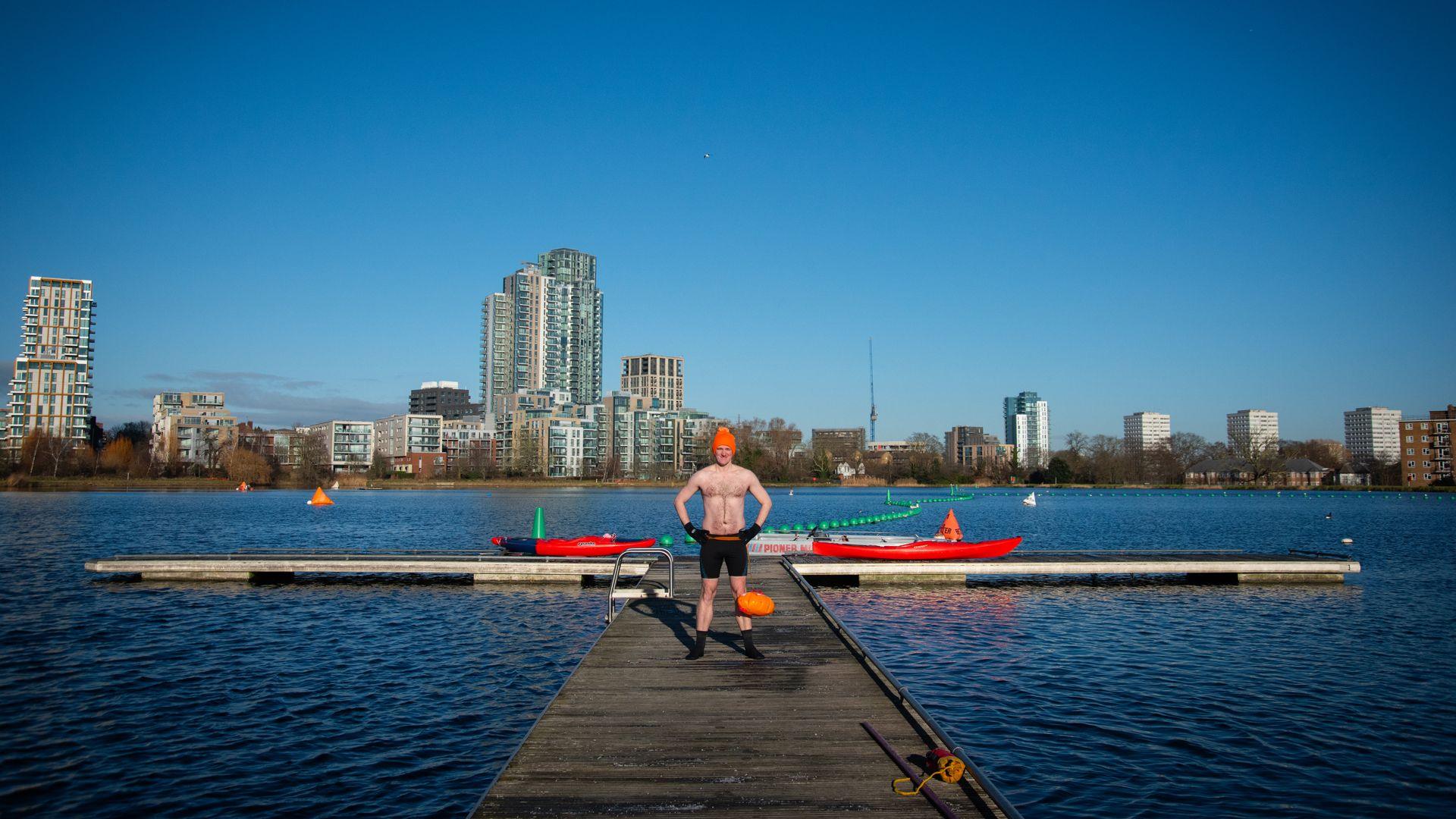 James stands on a ramp out across the west reservoir. He is wearing an orange bobble hat and swimming shorts as well as swimming socks. The sky is blue and there are high rise buildings in the distance, beyond the water's edge.