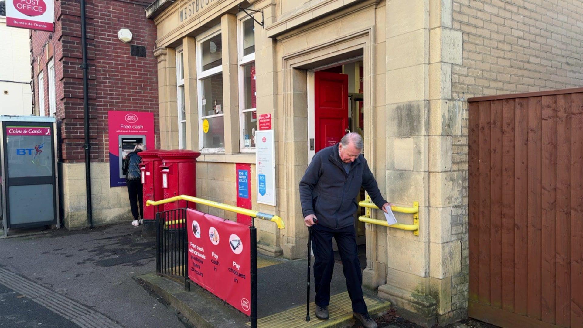 The exterior of Westbourne Post Office, it has two red letter boxes outside, next to a cash point which has a woman, with blonde hair wearing all black and trainers withdrawing money. There is a Post Office sign attached to the side of the building. Out the front, yellow side rails as support and a small ramp to get into the building, which has a red front door. There is an elderly man, using a black walking stick and wearing dark blue jeans and a dark grey fleece walking down the step outside the post office.