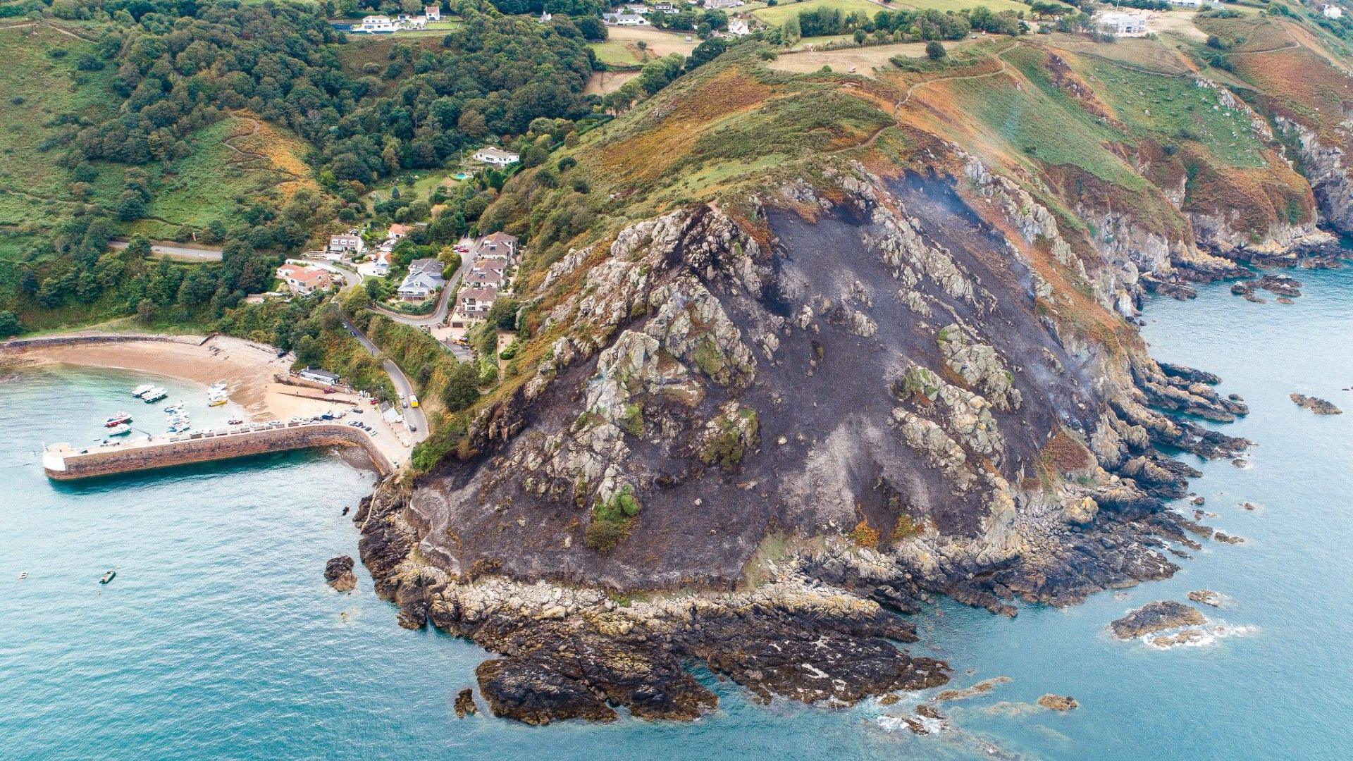 Aerial shot of a wildfire at Bonne Nuit. Smoke is billowing off the open area which has been damaged in the blaze. Water is surrounding the edge of the island.