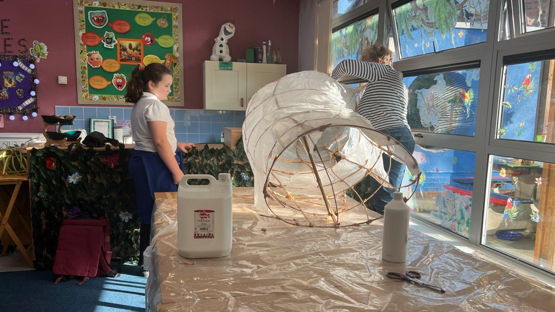 Child looking at teacher holding a large sculpture made out of sticks and glued paper.