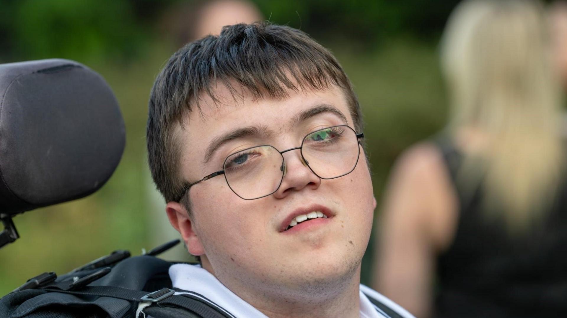 Mr Mayhew-Kemp smiles at the camera. He has short brown hair which rests over his forehead. He also wear glasses. 