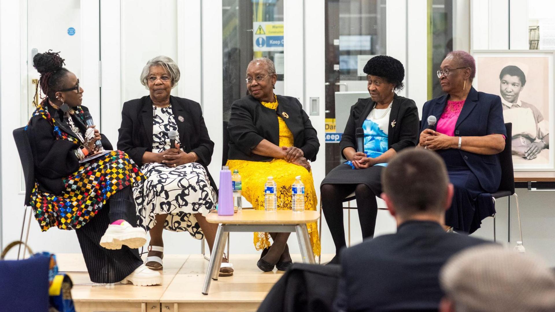 Five black women sit in a half circle with their hands folded in their laps. A table with bottles of water is positioned in the centre.