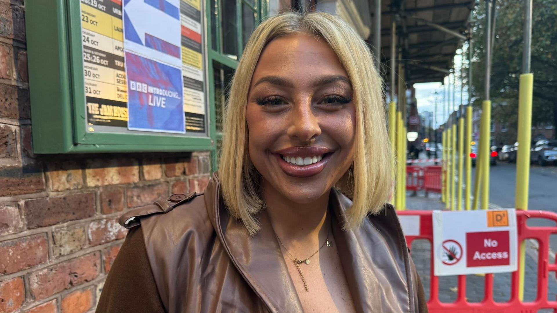 Maddie Juggins outside the BBC Radio 1Xtra Introducing gig smiling at the camera. She is wearing a brown leather jacket, and behind her are yellow construction poles with a red "no access" sign and barricade.