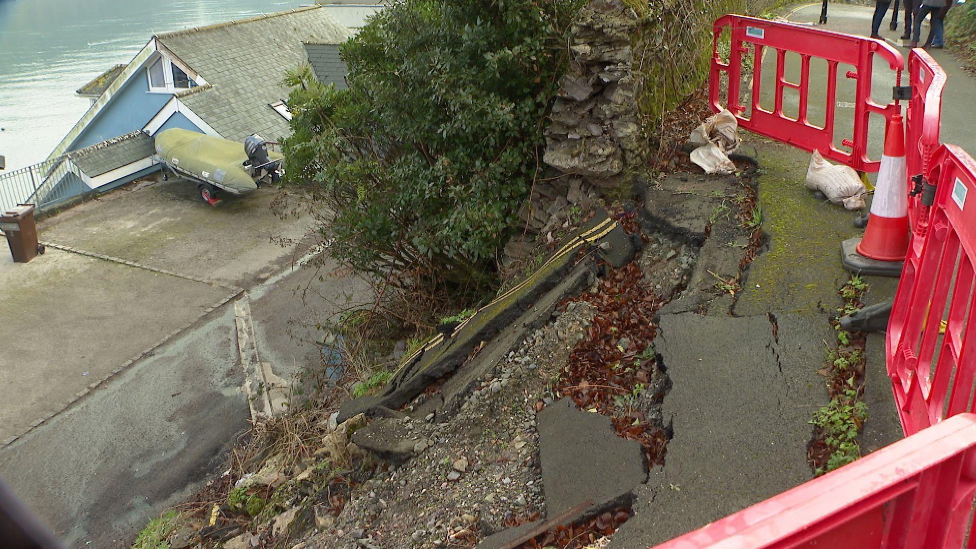 A collapsed wall which shows a large drop. The road at the top has cracks and the area is surrounded by red barriers. 