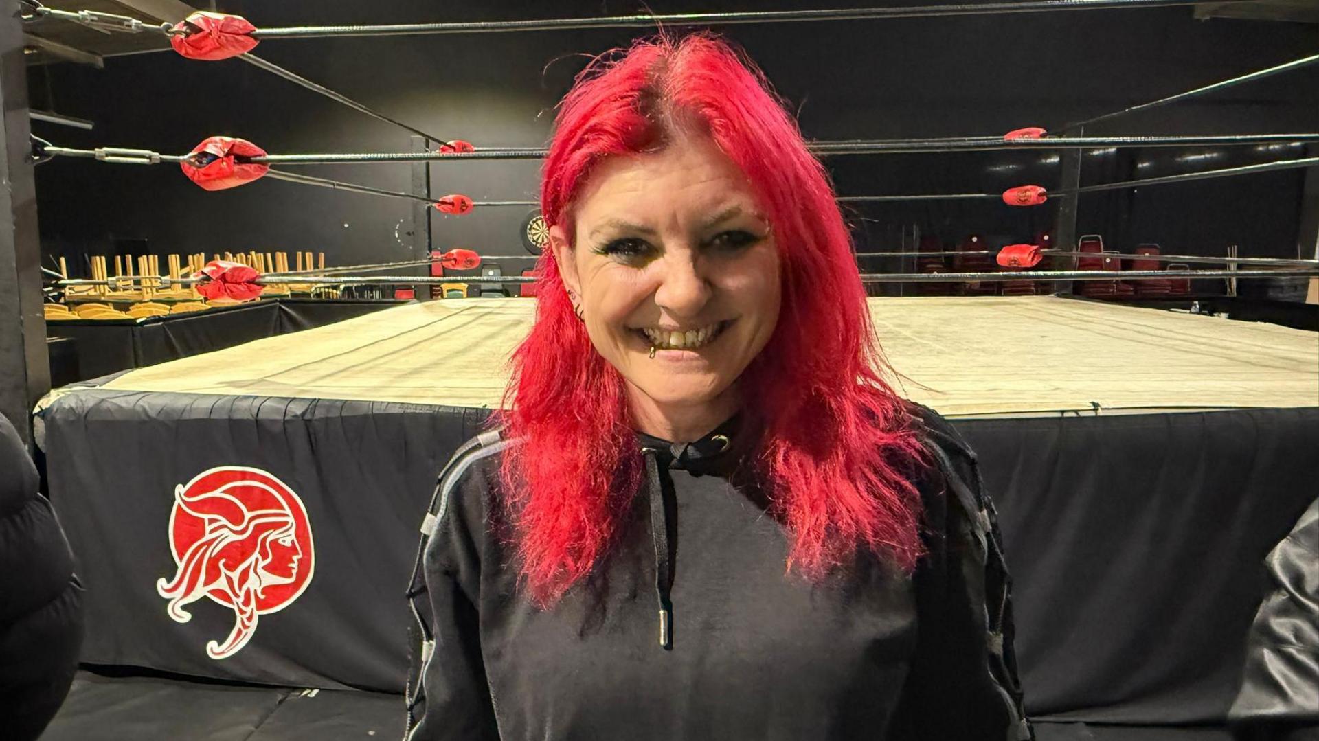 Wrestler Saraya stood beside a ring. She has long hair dyed red and is wearing a black hoodie. She is smiling at the camera.