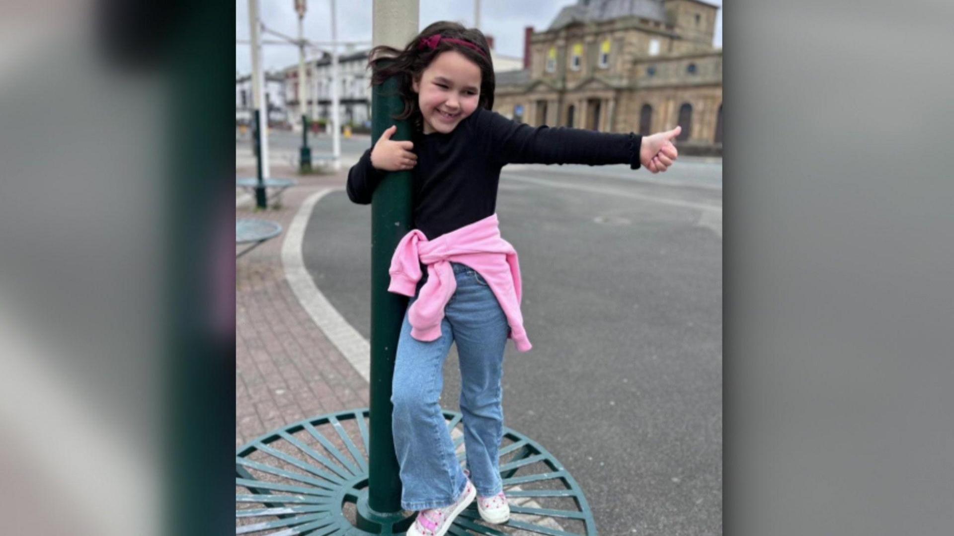 Bebe King, wearing jeans, a black top and a pink jumper around her waste, smiles and gives a thumbs up as she whirls around a lampost