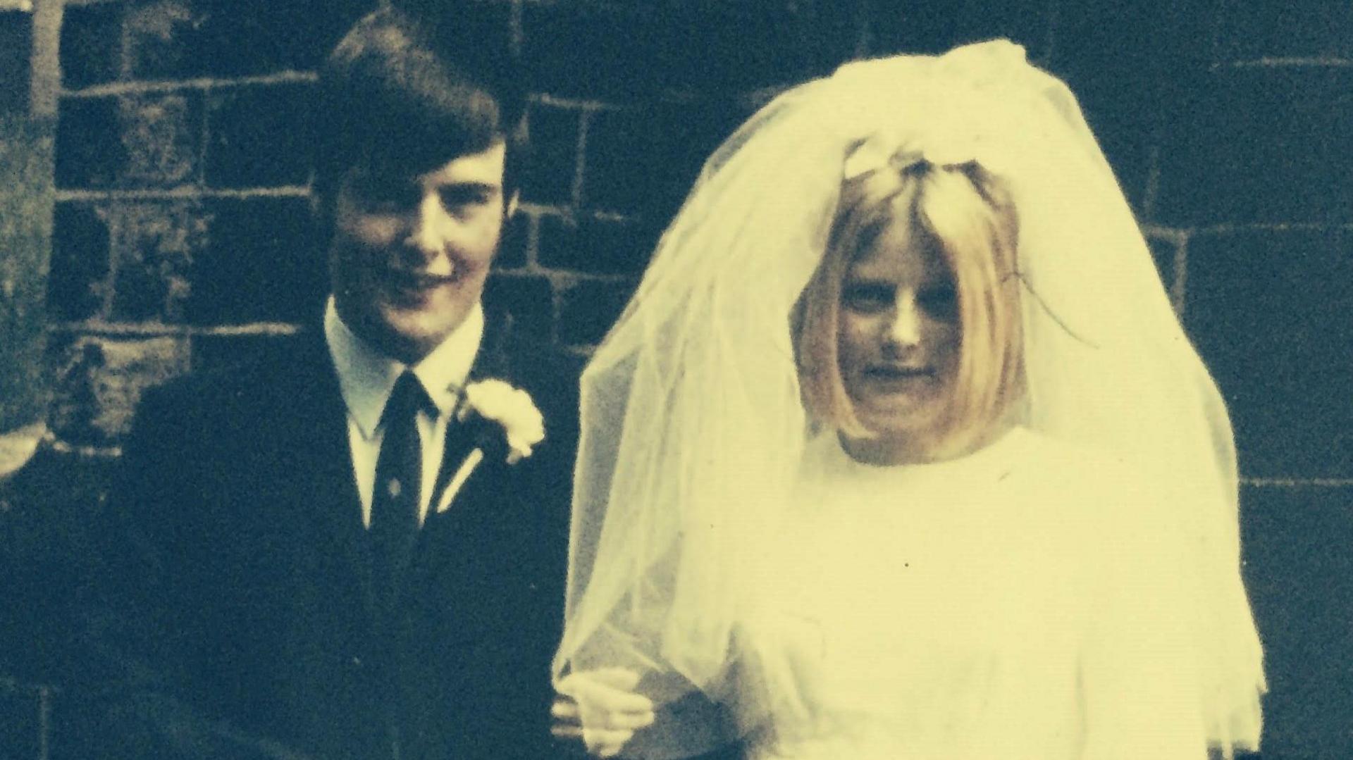 John on the left in a black suit and tie with a white flower on his jacket and Pat in a white wedding dress and veil